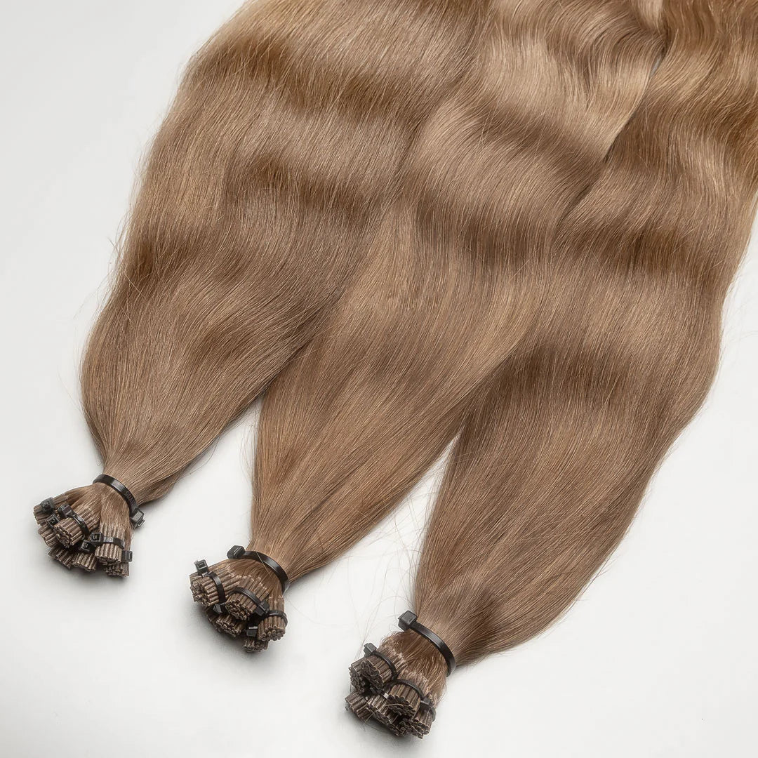 A photo of I-tip hair extension strands with a golden dark blonde color, secured with small beads.