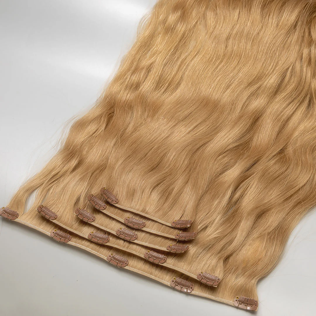 A section of clip-in, dark blonde hair extensions attached to small metal clips, laid out on a white surface.